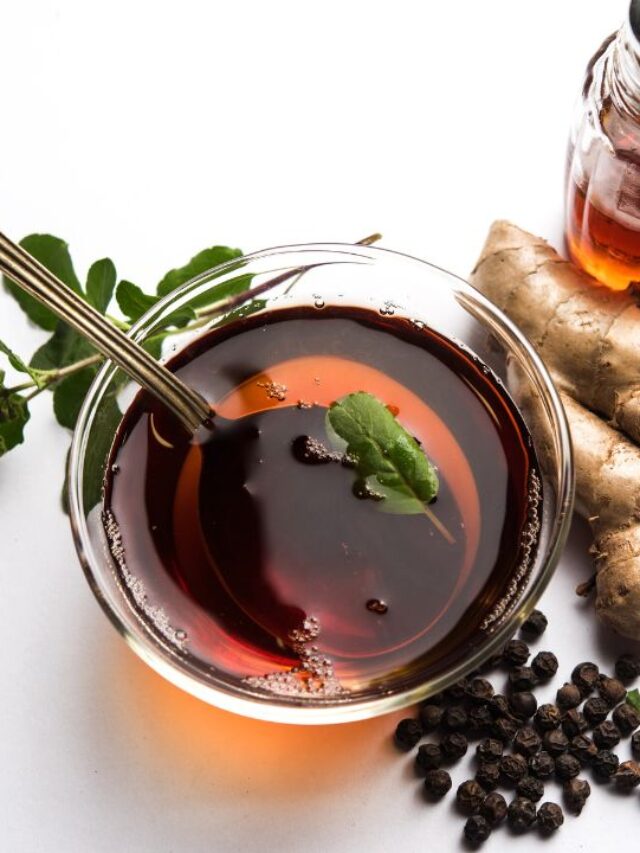 kadha in cup with spoon. Beside the cup ginger, lemon slice, basil leaves, clove and black peppers on the surface.