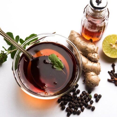 kadha in cup with spoon. Beside the cup ginger, lemon slice, basil leaves, clove and black peppers on the surface.