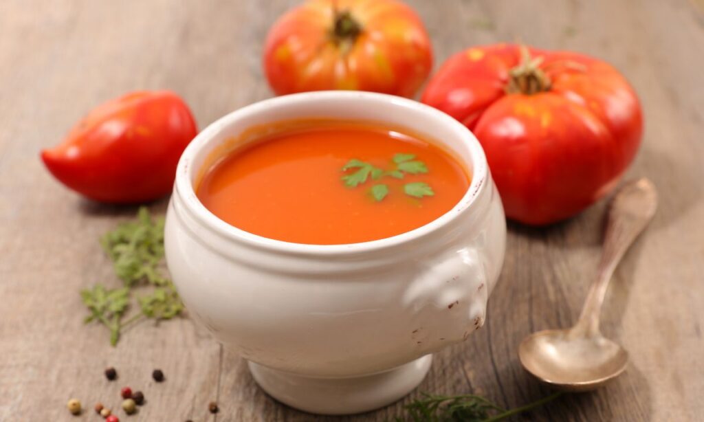 tomato soup in white color bowl, beside the bowl spoon and tomatoes on the surface.