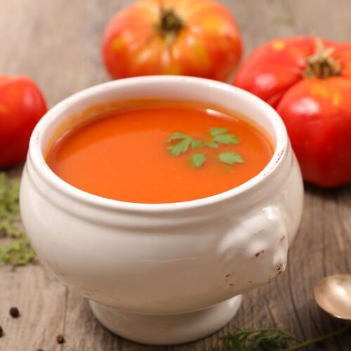 tomato soup in white color bowl, beside the bowl spoon and tomatoes on the surface.