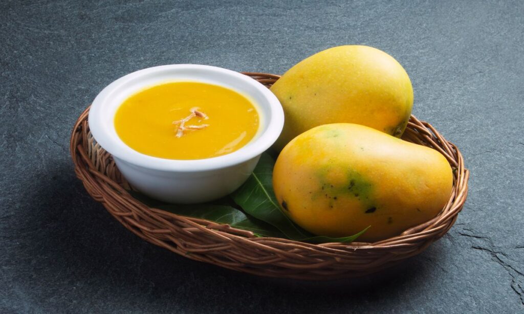 aamras in white color bowl. beside the bowl of aamras two mangoes.