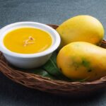 aamras in white color bowl. beside the bowl of aamras two mangoes.